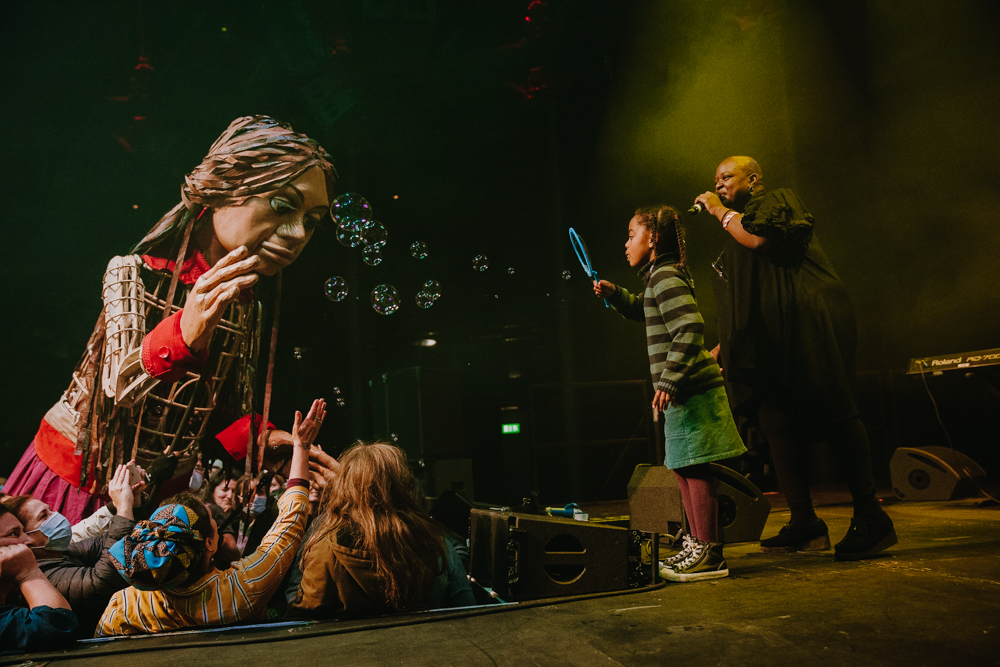 A little girl onstage blows bubbles towards Little Amal, a 3.5 metre high puppet, whilst a ESKA sings on stage. The crowd reaches for the bubbles.