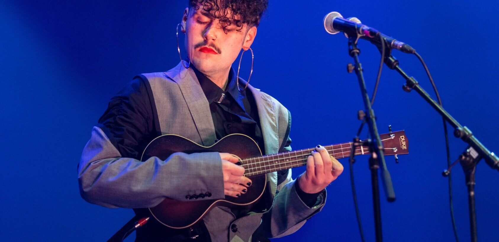 Luca Manning performs on stage behind a microphone with their eyes closed and playing the ukulele.