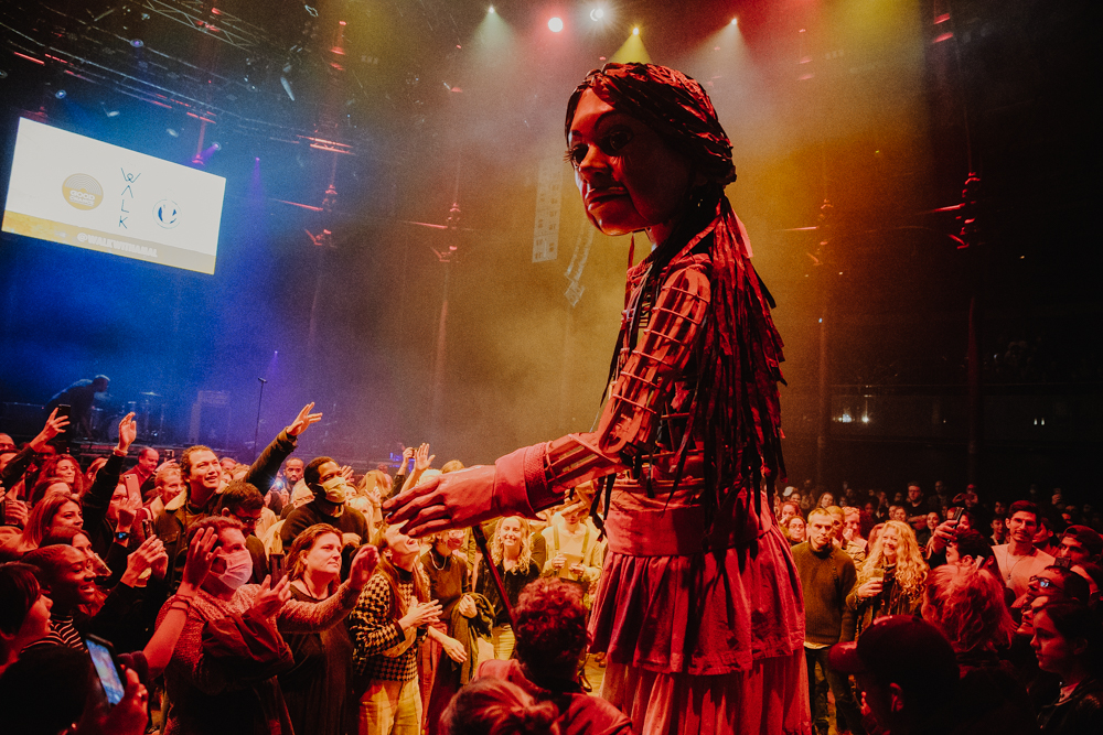 Little Amal stands in the middle of a crowd of people who are all looking with wonder and reaching out to her.