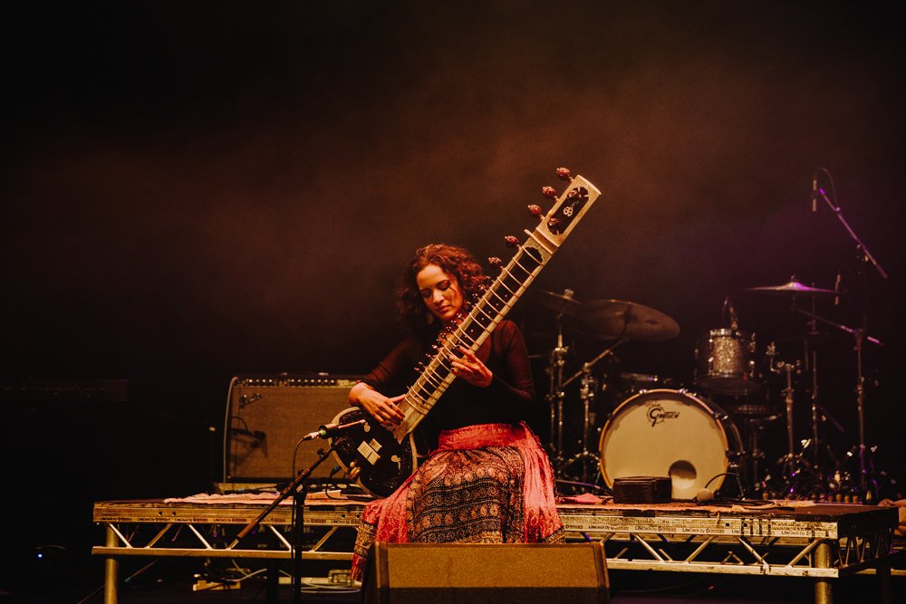 Anoushka Shankar plays the sitar on stage with her eyes closed sitting against the backdrop of other musical instruments.