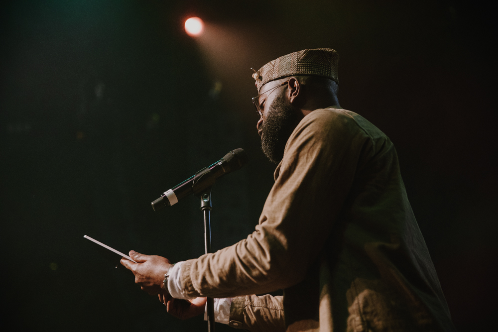 Inua Ellams stands in front of a microphone holding out a poem that he is reading to the audience.