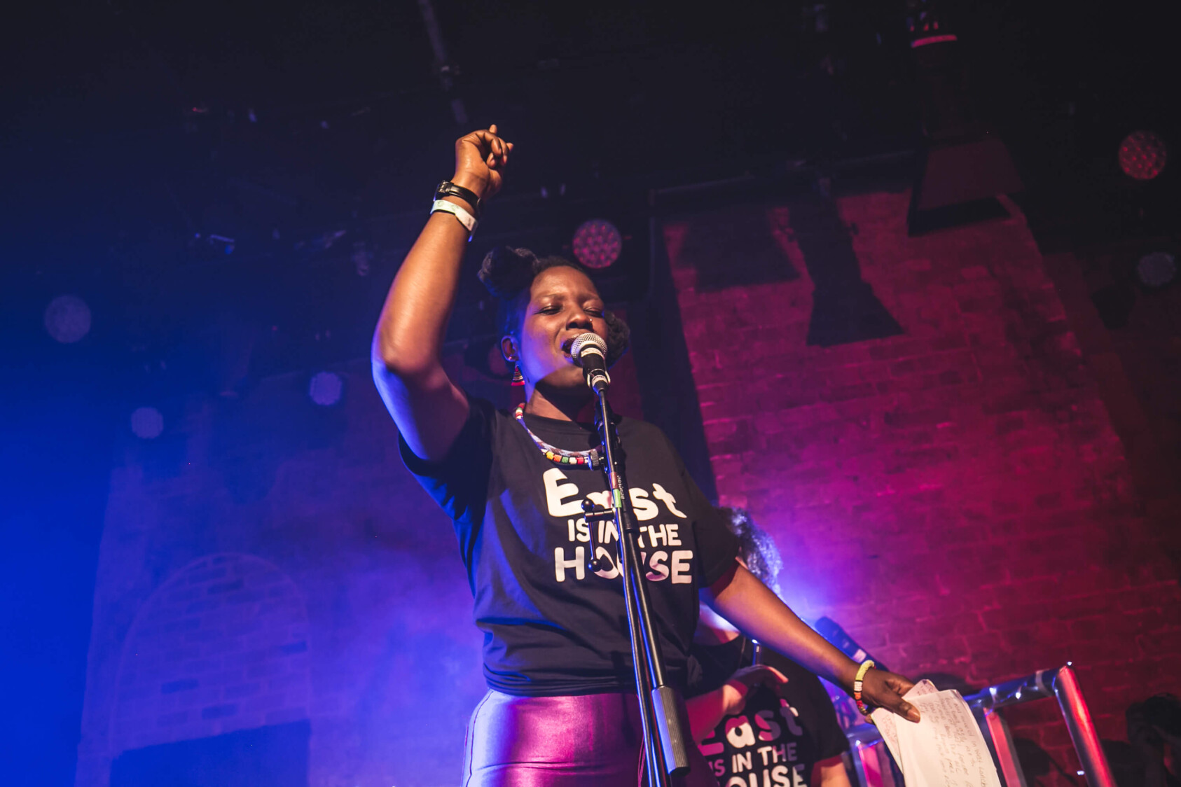 Poet Wunmi Onibudo performs on stage at Boxed In. She has one hand raised above her head and hold a book in another. She is wearing a black t-shirt.