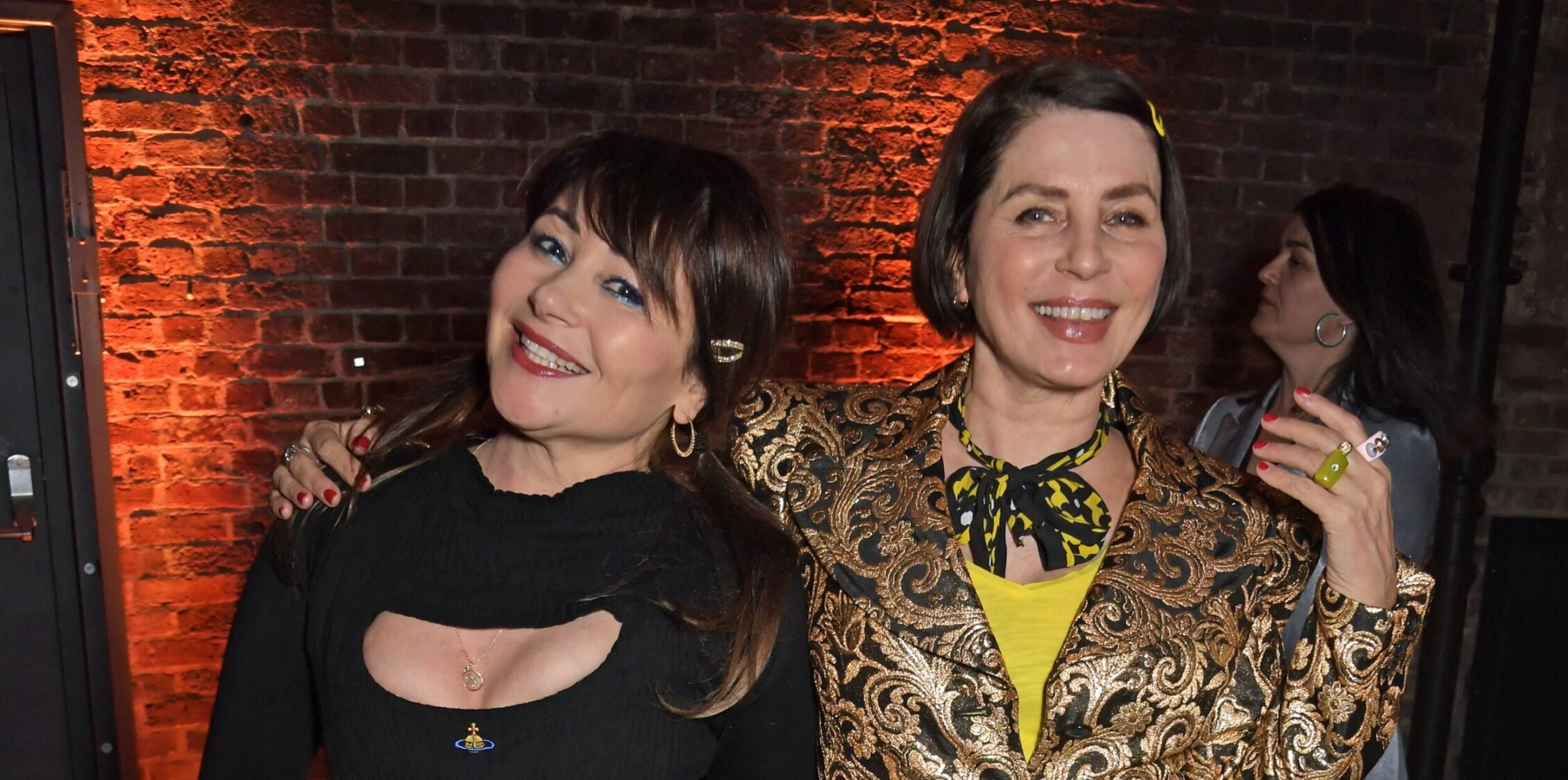 Frances Rufelle and Sadie Frost stand with their arms around each other, smiling to the camera, with the brick wall of the Roundhouse in the background.