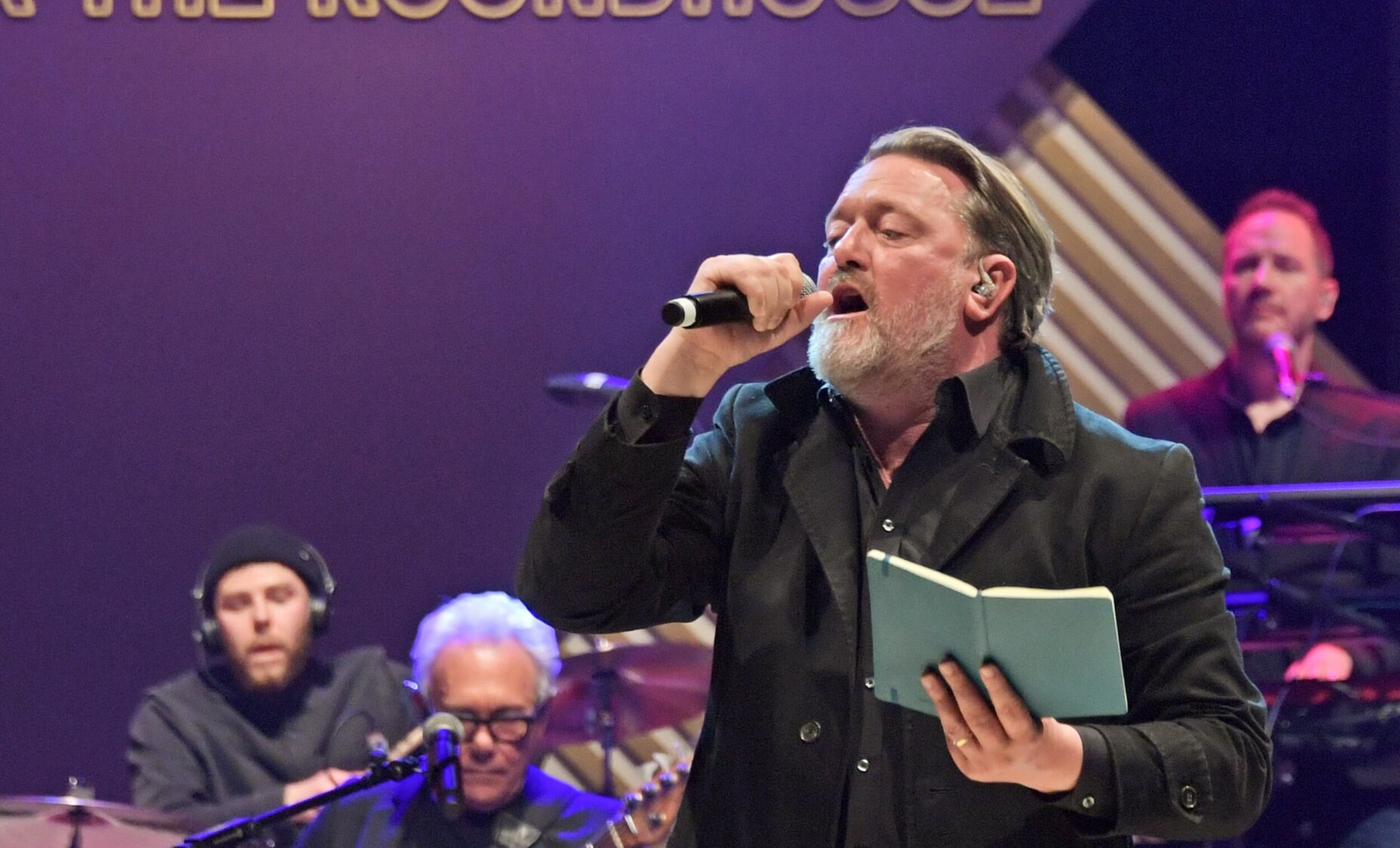 Guy Garvey sings to the crowd whilst holding a notebook in his hands.