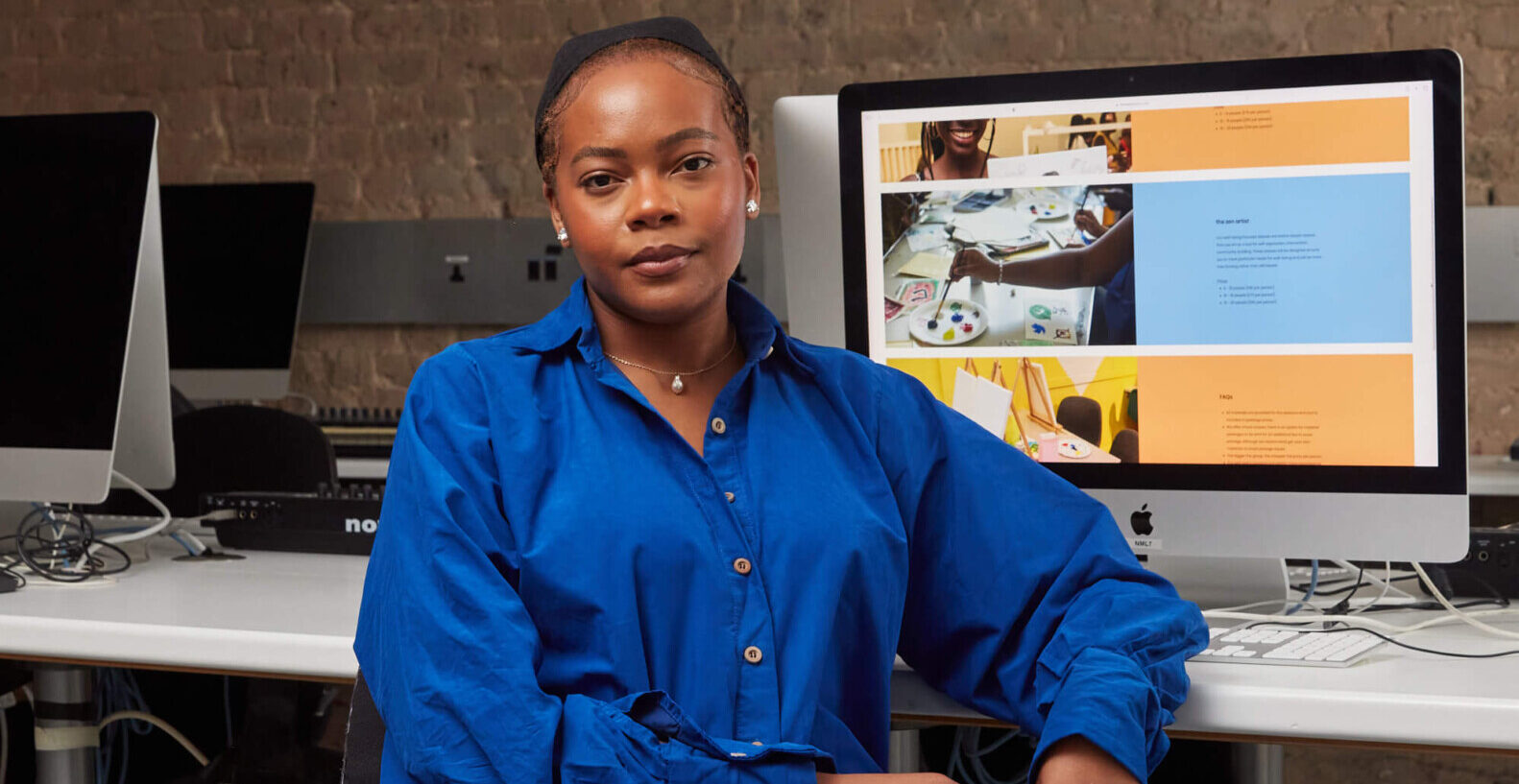 A young woman dressed in blue sits at a desk with a computer screen behind her