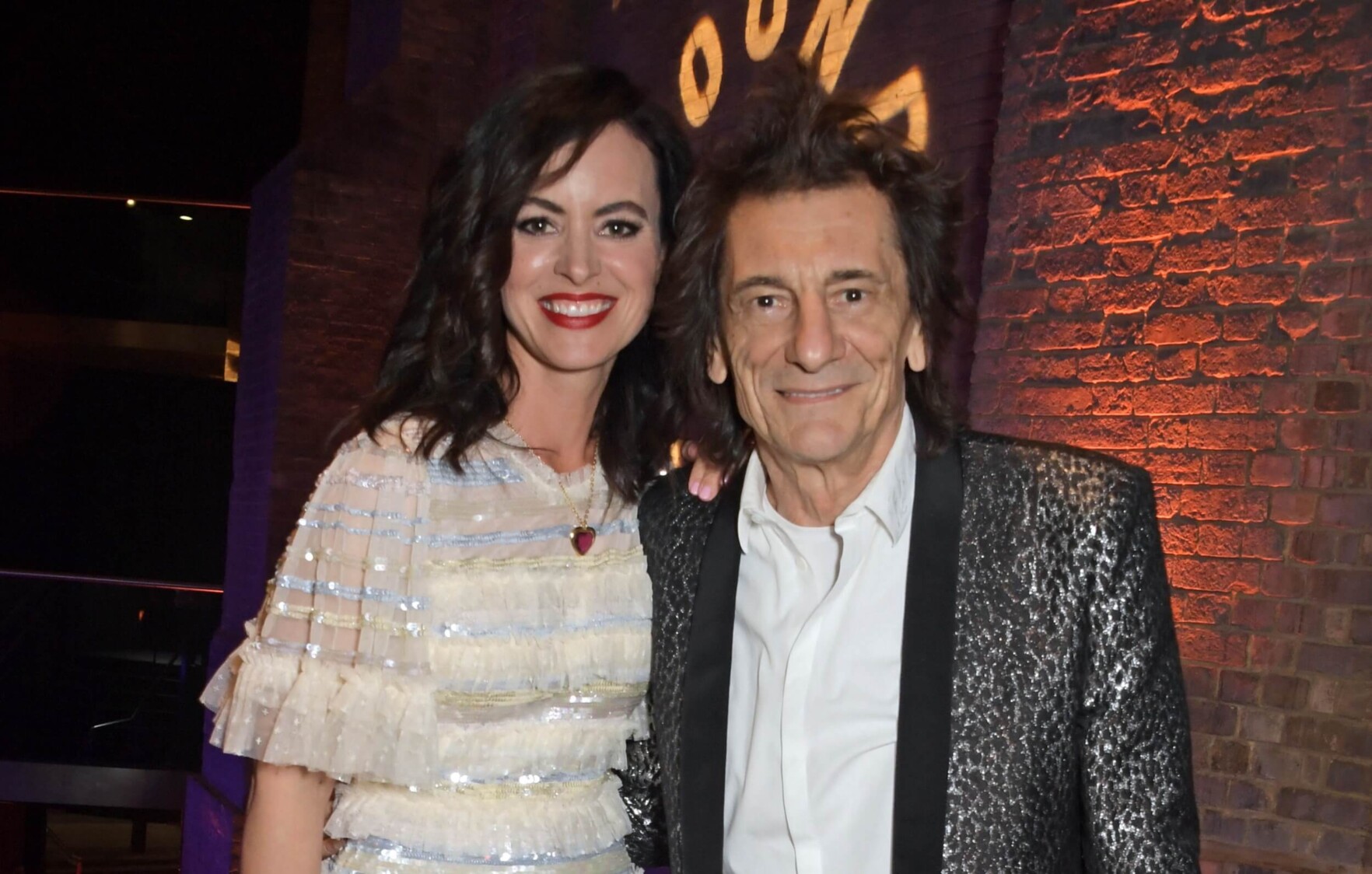 Sally Wood and Ronnie Wood stand next to each other, smiling to the camera, with the brick wall of the Roundhouse in the background.