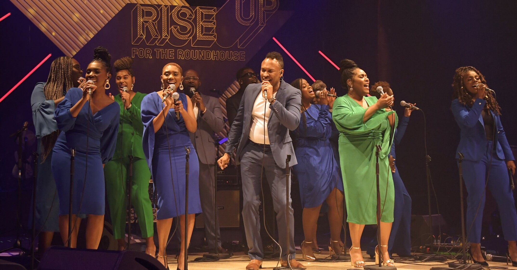 Twelve members of The Kingdom Choir are singing on stage together whilst smiling and looking out to the audience.