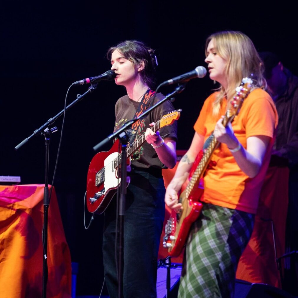 Two members of the band Goat Girl perform on stage with guitars.