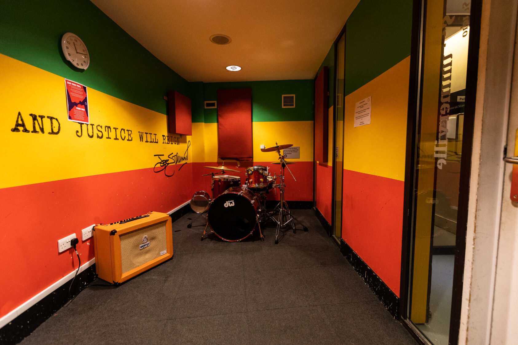 inside of a room painted red, yellow and green stripes. There is an orange amp on the left and a drum kit at the far end of the room