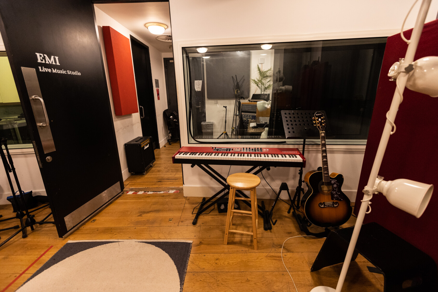 The interior of the recording studios, there is a guitar, jeyboard and stool. Visible through the window is a mixing desk