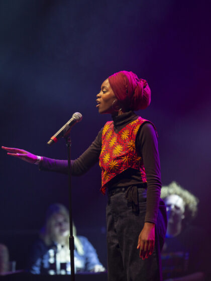 A woman in an orange and red vest wearing a red head wrap speaks into a microphone with her right hand extended in front of her