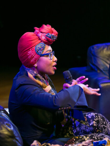 A woman in a red headwrap and glasses sitting down speaking and holding a mircrophone