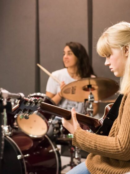 A young woman with blonde hair plays the guitar while a woman with mid-length dark hair plays the drums in the background