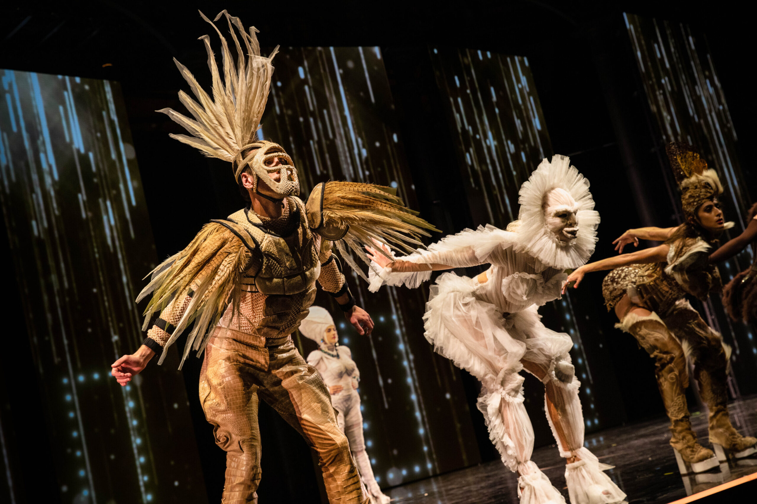 Dancers on stage wearing lace and feather outfits in front of a digital screen that looks like falling rain