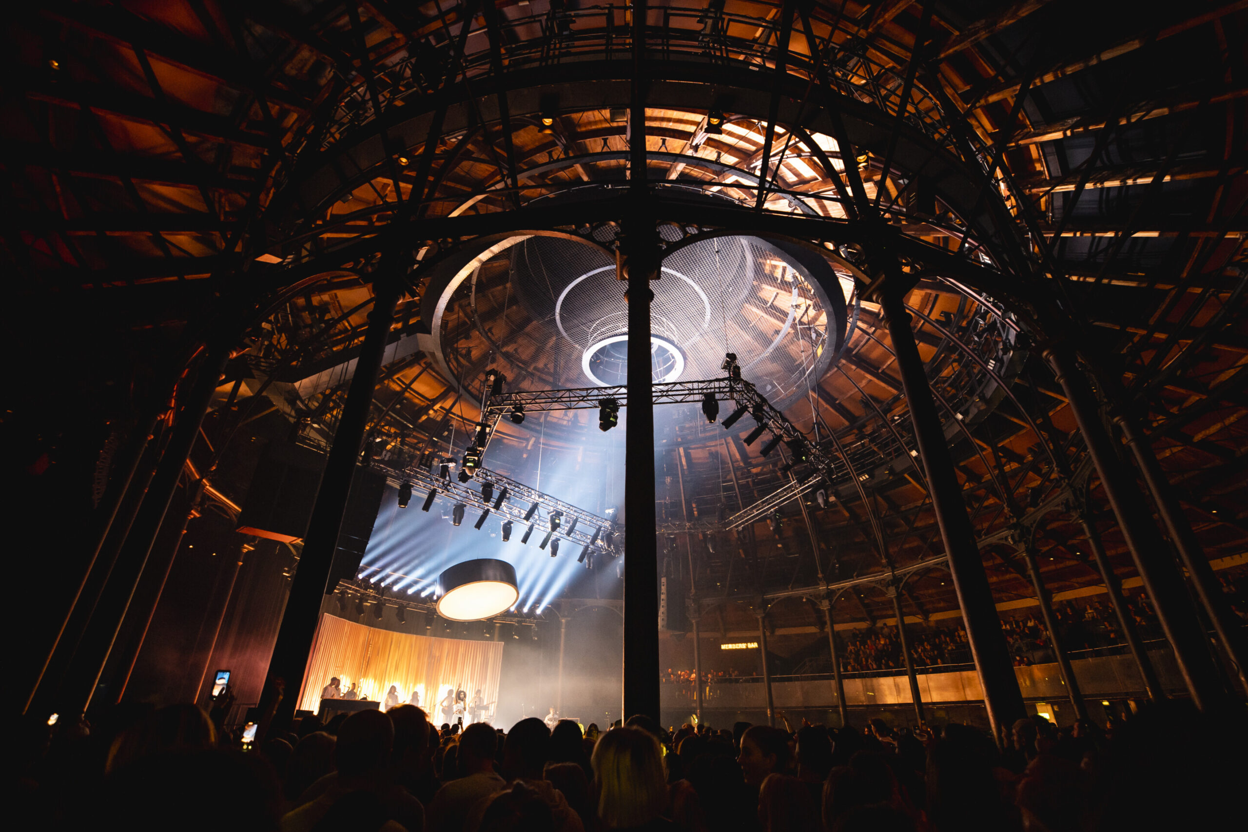 A large crowd stands in the main auditorium of the Roundhouse and you can see the performer on stage and the architecture of the roof