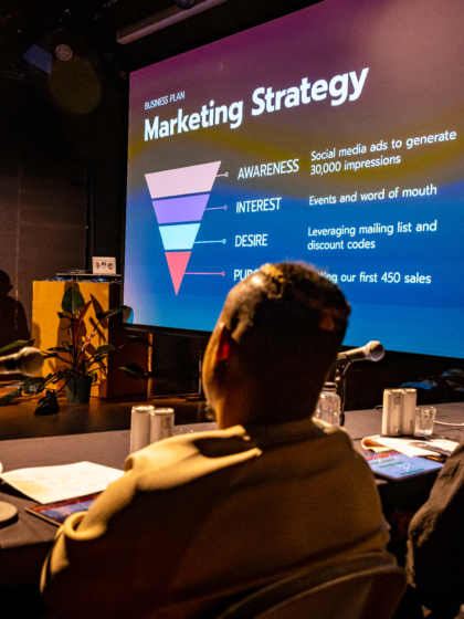 Two presenters on a stage with a screen behind them displaying a marketing strategy funnel. The screen shows a multi-level funnel labeled 'AWARENESS', 'INTEREST', 'DESIRE', and 'PURCHASE', with corresponding marketing tactics listed next to each level. In the foreground, the backs of a few audience members are visible.