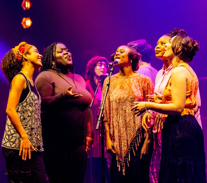 A group of young people singing joyfully around a microphone on stage at the Roundhouse. Performing the songs of Joni Mitchell.