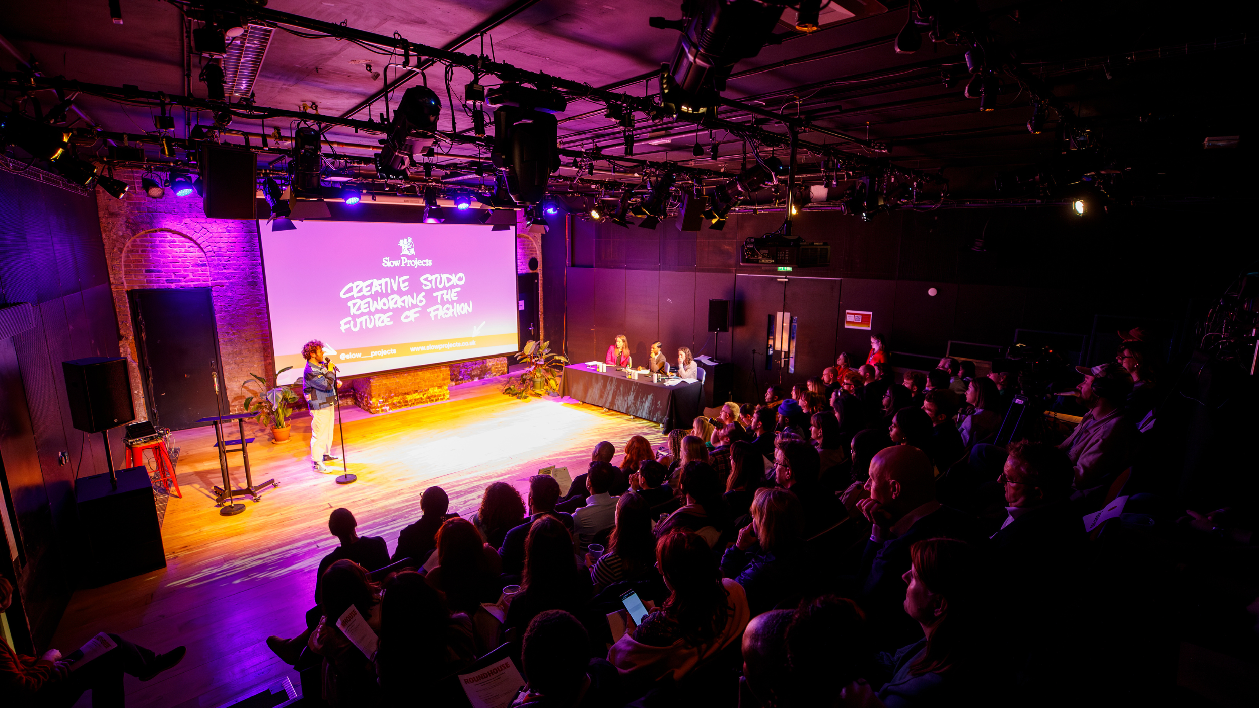 an audience of roughly 50 people watching one person pitch their presentation to a panel of three judges