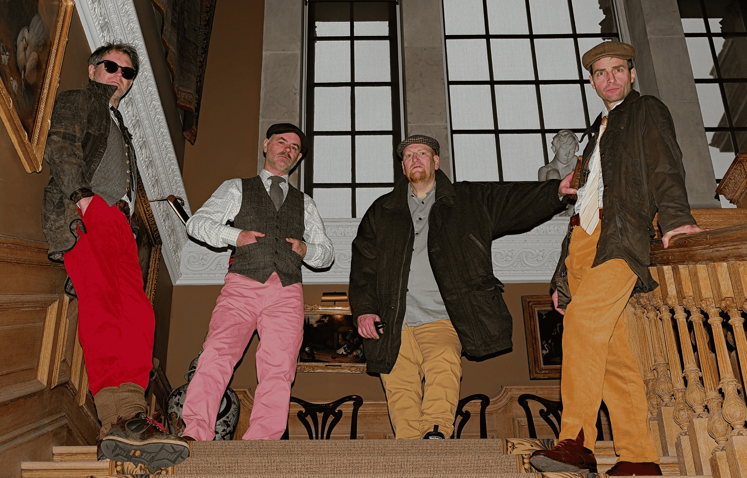 Four members of The Beta Band posing on an elegant staircase in a historic building. The musicians wear an eclectic mix of vintage-inspired clothing and flat caps.