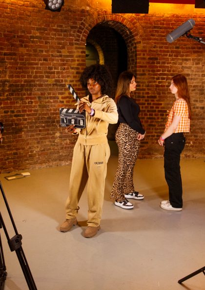A film crew captures a scene with two actors standing face-to-face, while a clapperboard operator, camerawoman, and boom mic operator work around them in a brick-walled studio.
