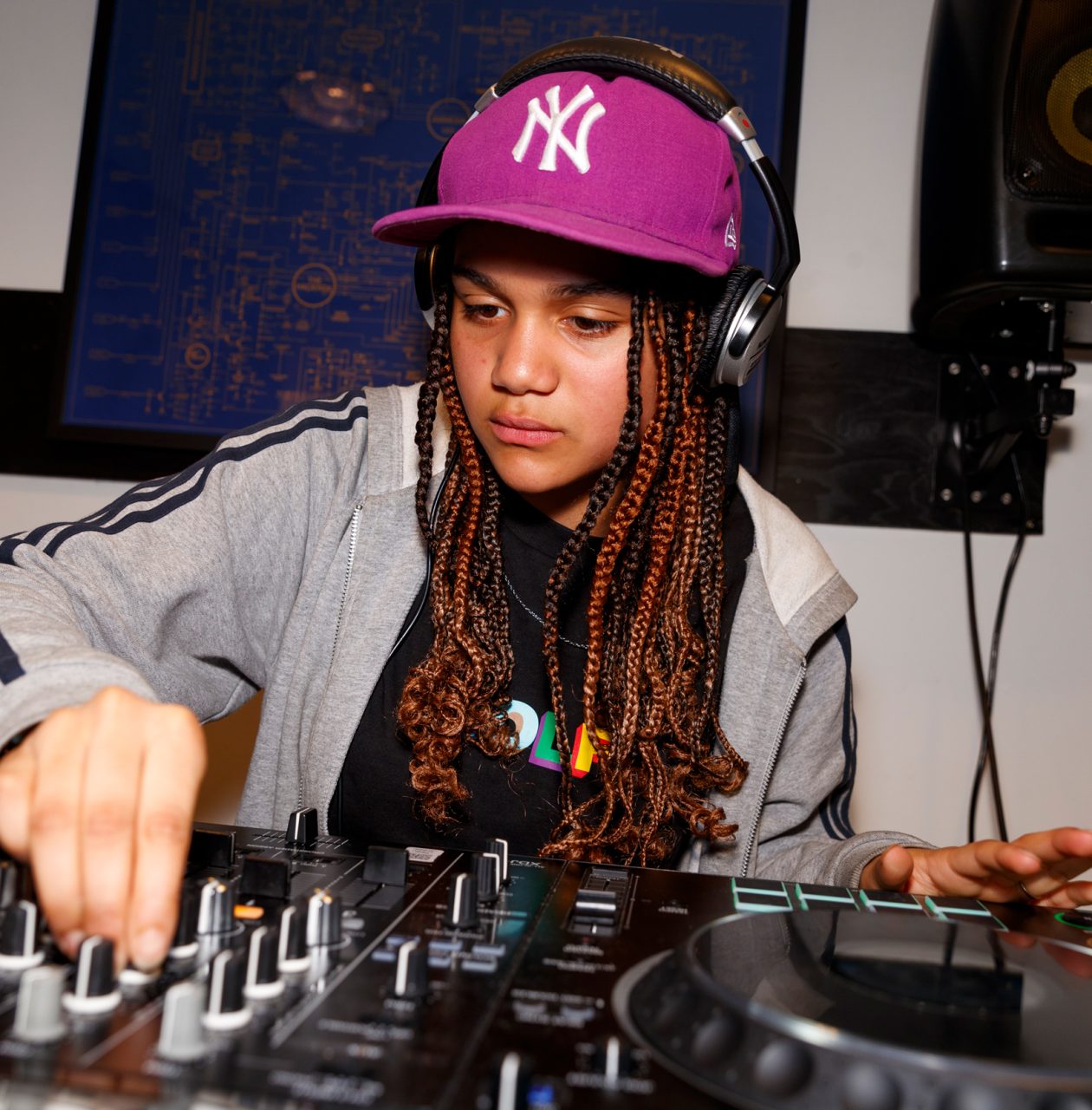 Young DJ with long braids, wearing a purple Yankees cap and headphones, adjusts knobs on a mixing console while performing in a studio.