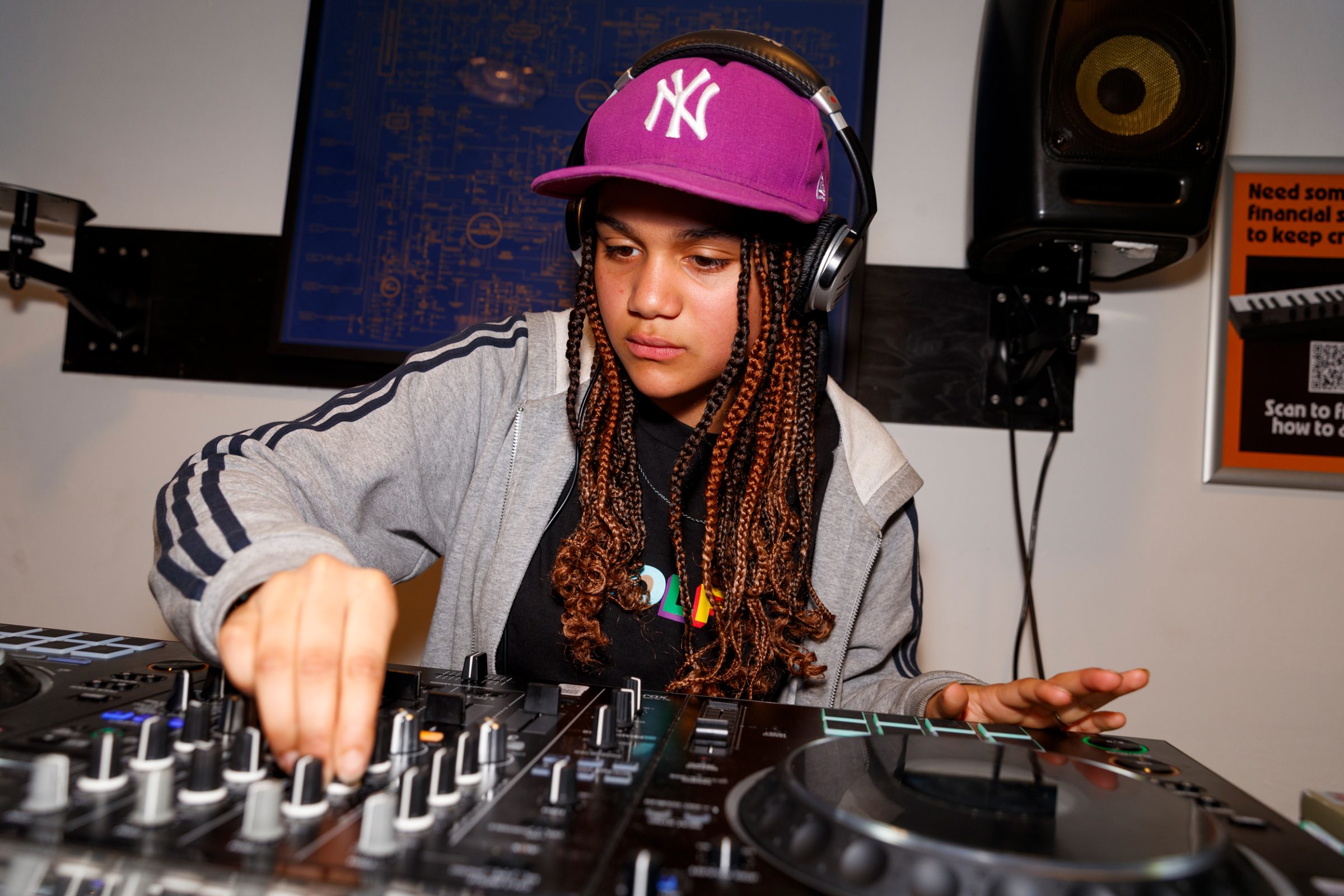 Young DJ with long braids, wearing a purple Yankees cap and headphones, adjusts knobs on a mixing console while performing in a studio.