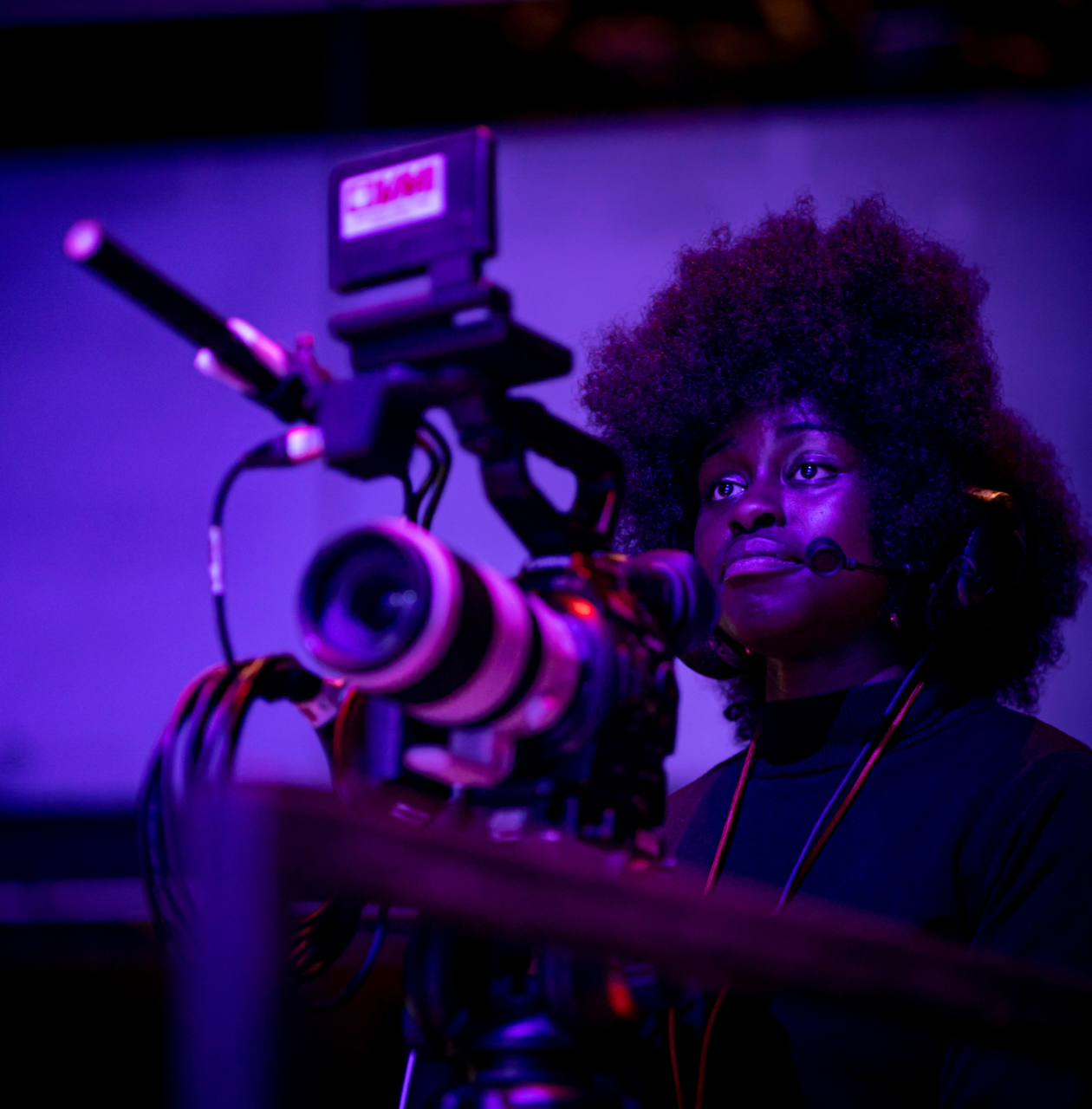 A camera operator wearing a headset films under dramatic purple lighting, focused intently behind a professional video camera.