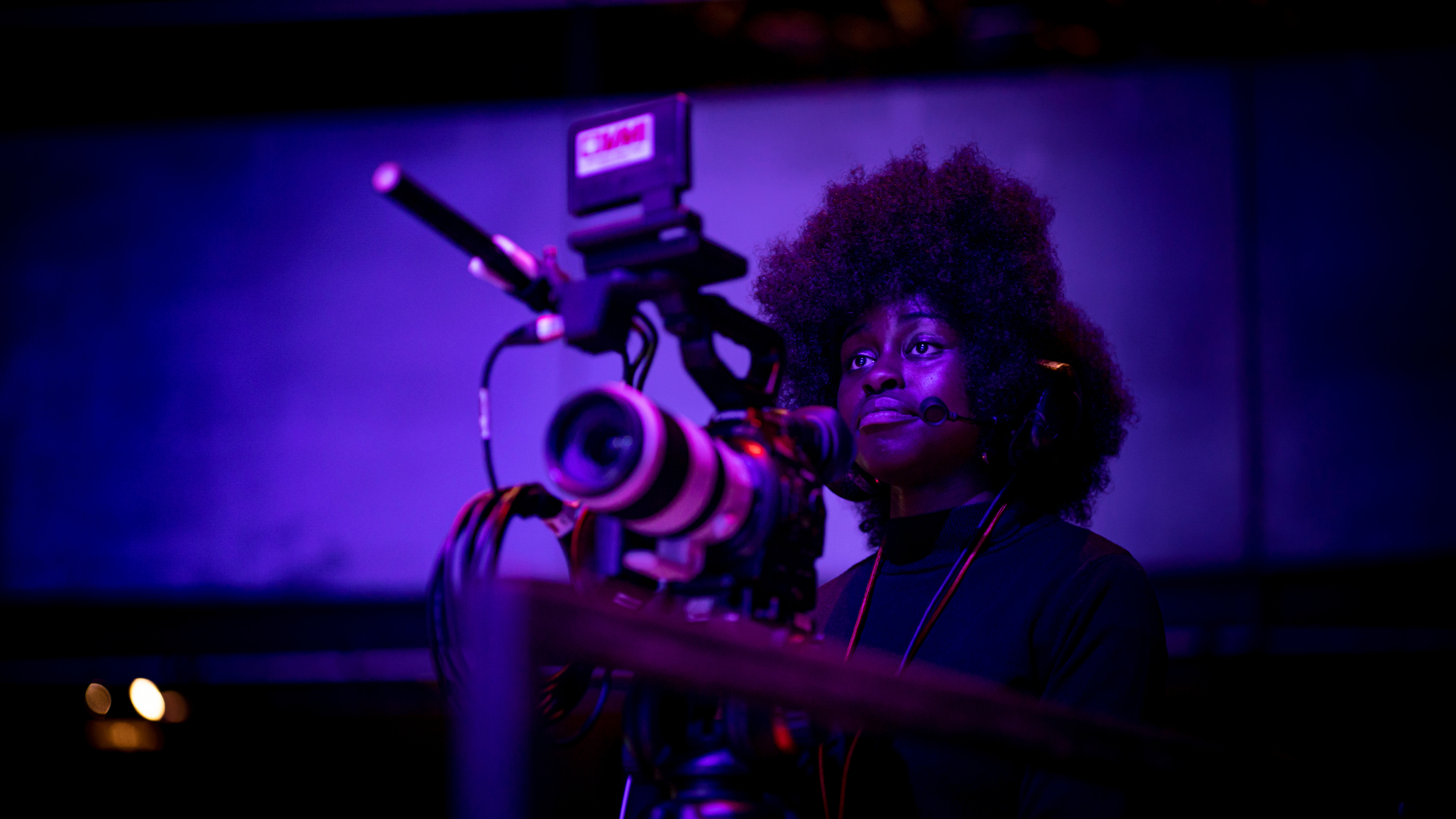 A camera operator wearing a headset films under dramatic purple lighting, focused intently behind a professional video camera.