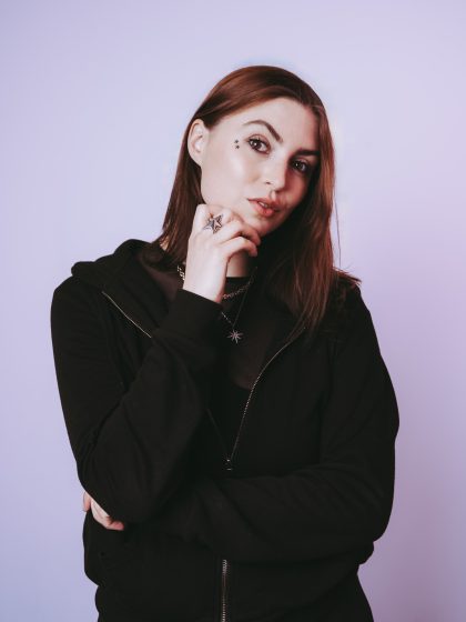 Young person with pale skin, long brunette hair wearing a black hoodie