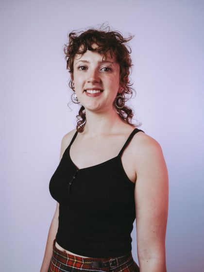 young person with pale skin, short curly brown hair wearing a black vest top