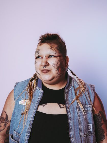 young person with short hair and facial tattoos, wearing a ripped black top and denim shirt