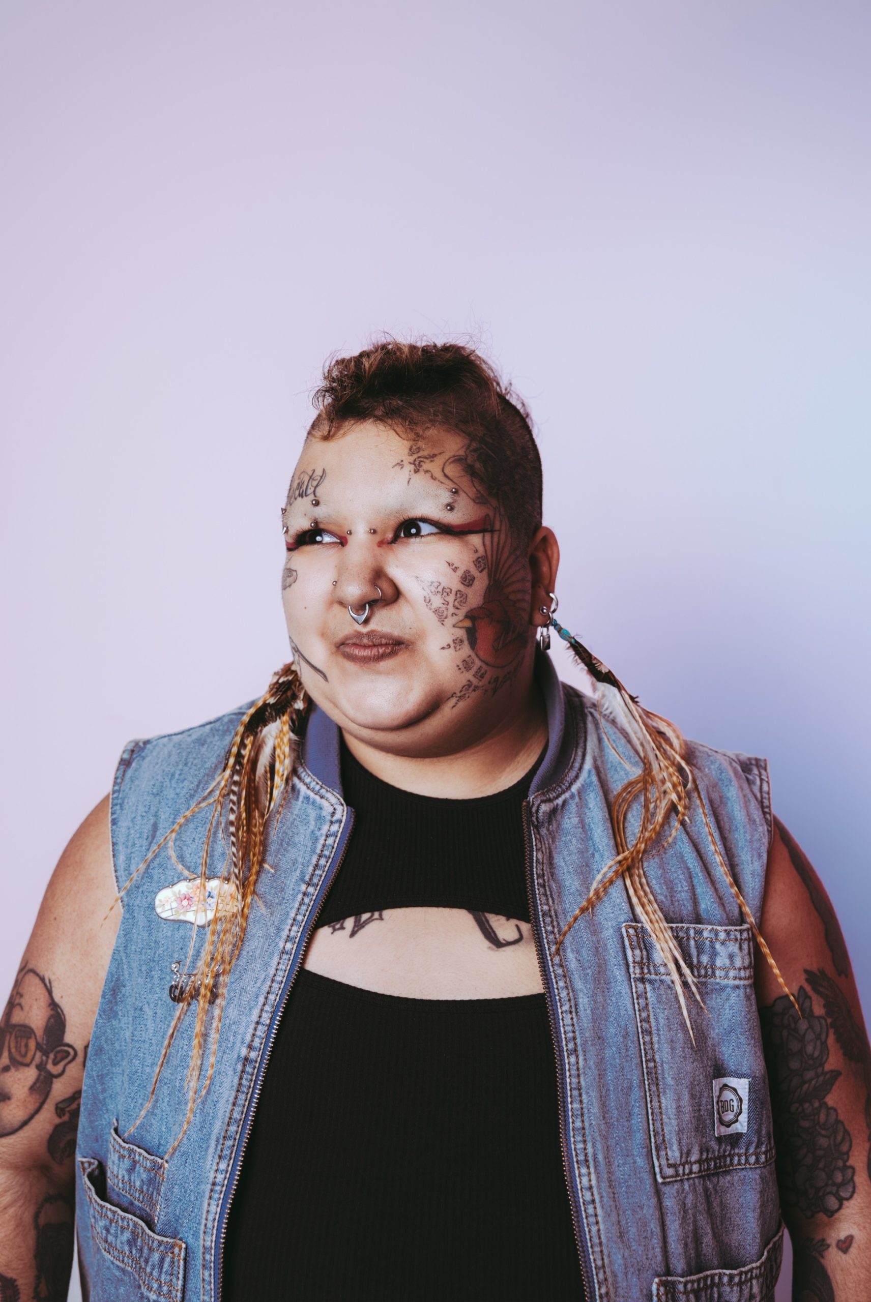 young person with short hair and facial tattoos, wearing a ripped black top and denim shirt