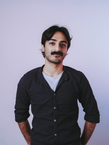 young person with dark skin, black hair and a mustache, wearing a long sleeve black jumper