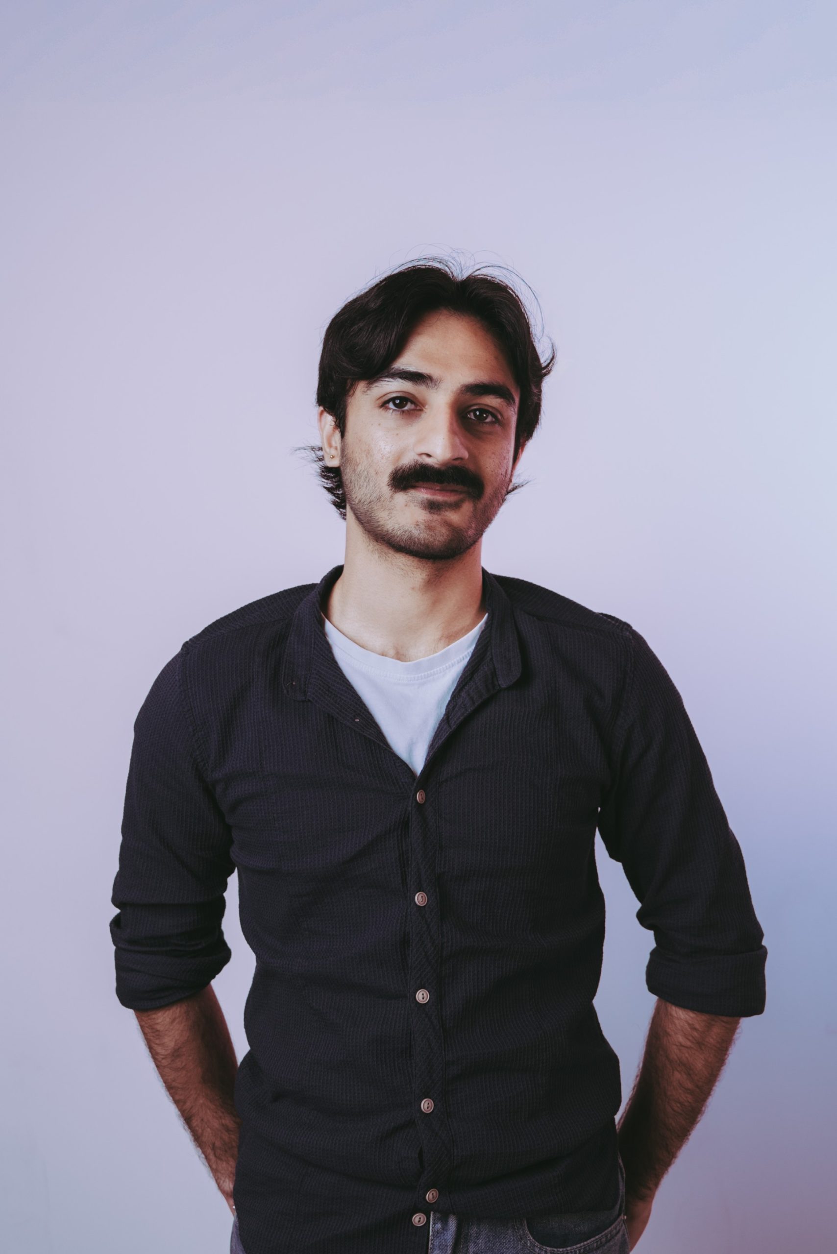 young person with dark skin, black hair and a mustache, wearing a long sleeve black jumper