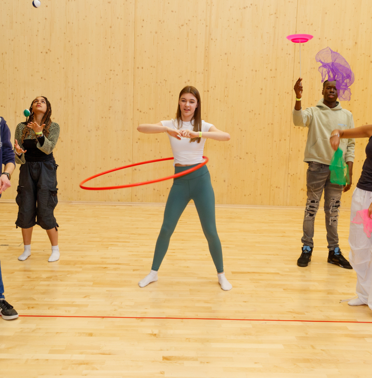 Five young people practicing circus skills indoors, including hula hooping, spinning plates, juggling scarves, and using a diabolo.