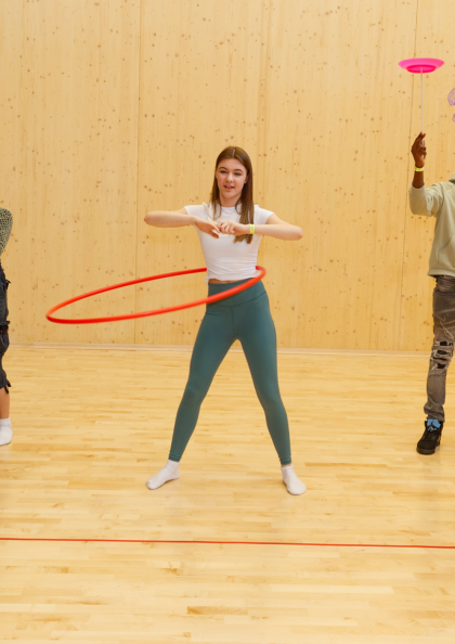 Five young people in a brightly lit room with a wooden floor practice various circus skills. From left to right: a male juggles with two diabolo sticks, a female juggles three balls, a female hula hoops with a red hoop, a male spins a plate on a stick and juggles a green item, and another female stands beside him holding a pink feathered object. Black curtains are visible on either side of the room