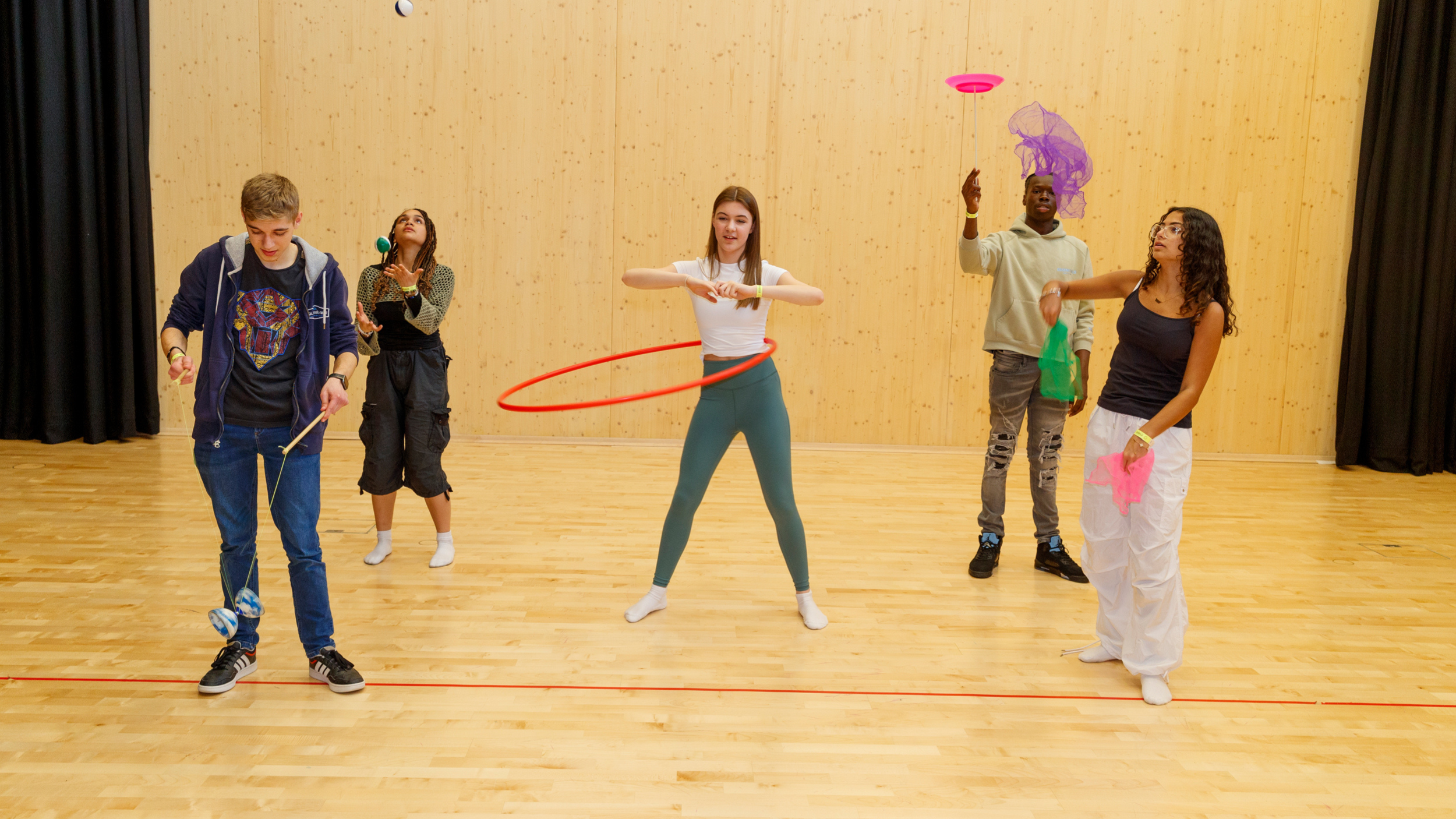 Five young people practicing circus skills indoors, including hula hooping, spinning plates, juggling scarves, and using a diabolo.