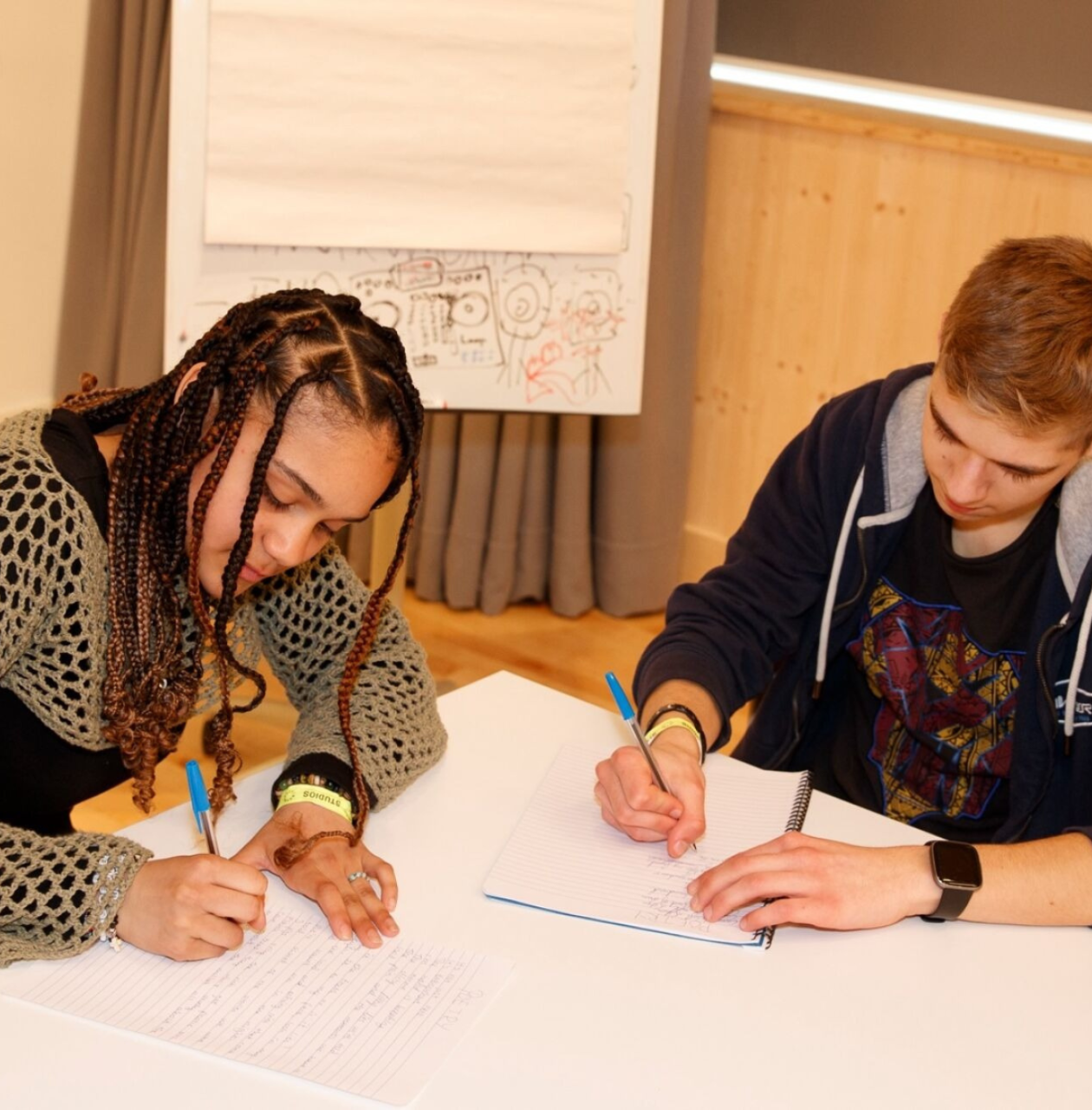 An image of two young people sitting at a table and writing in notebooks