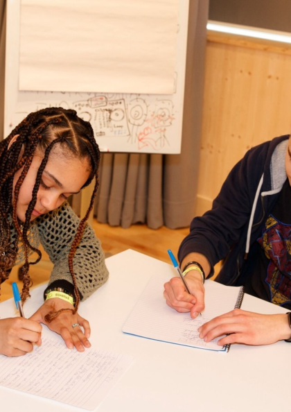 An image of two young people sitting at a table and writing in notebooks