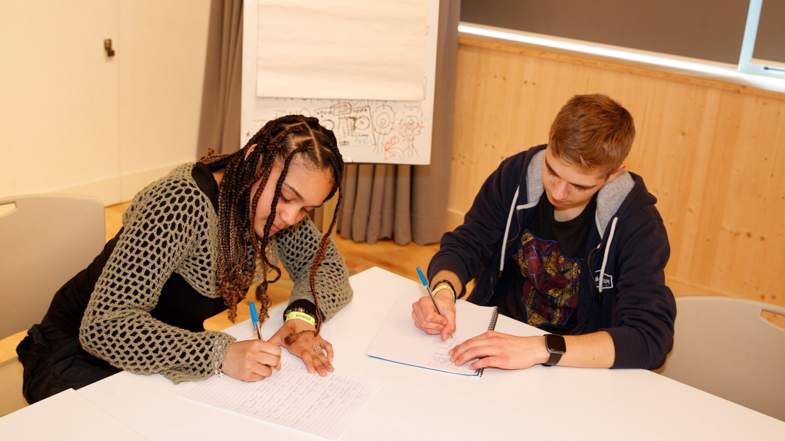 An image of two young people sitting at a table and writing in notebooks