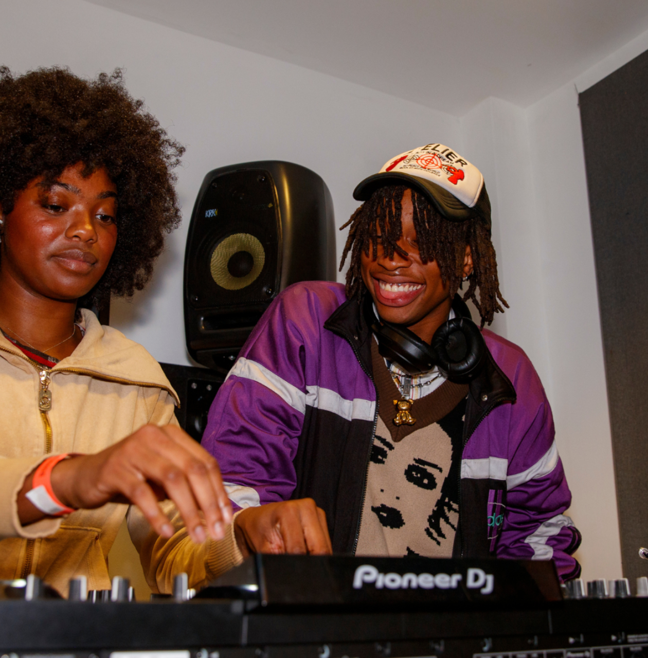 Two young DJs work together at a mixing console in a music studio, one focused and the other smiling.