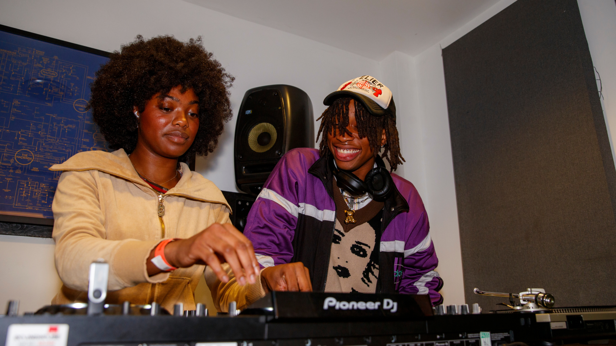Two young DJs work together at a mixing console in a music studio, one focused and the other smiling.