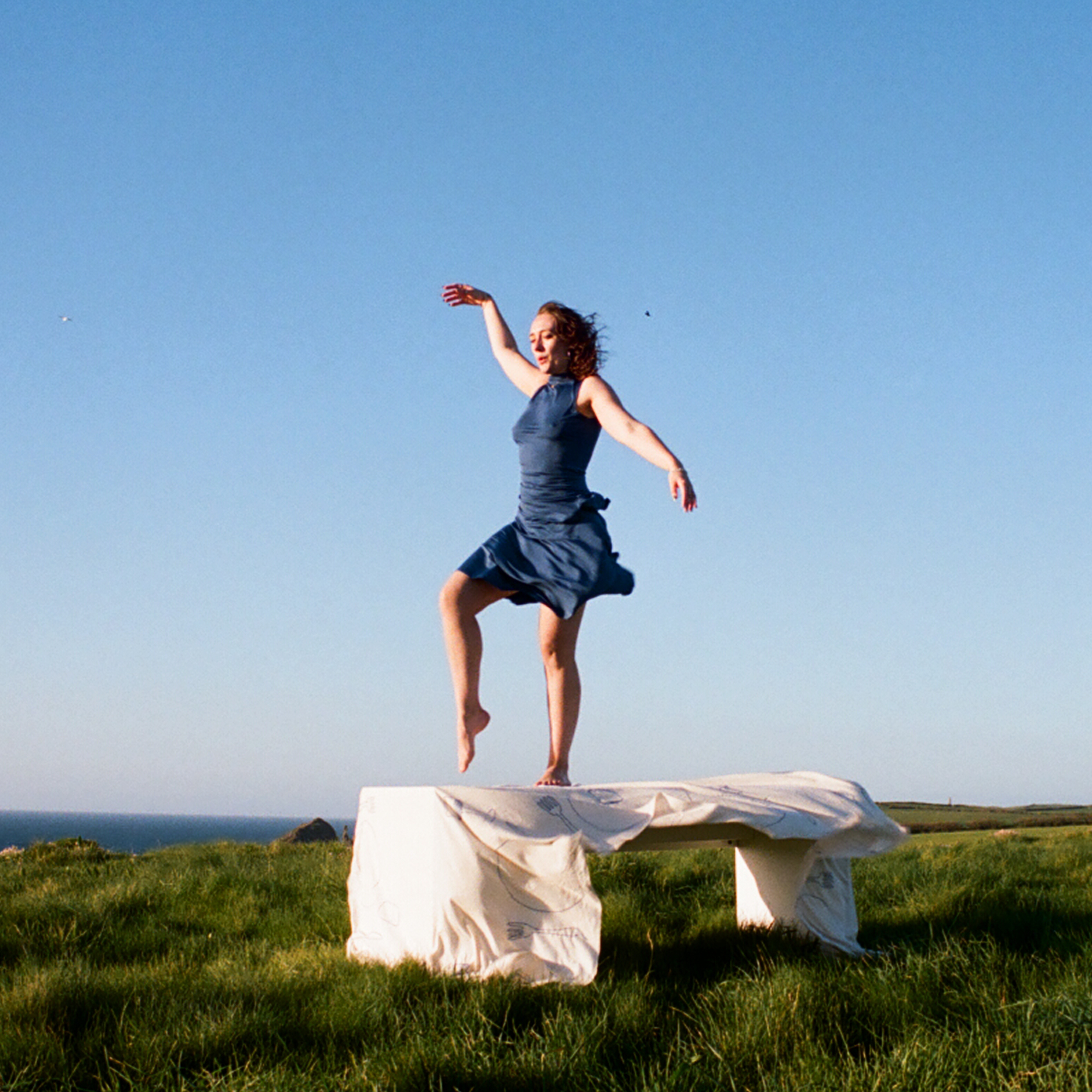 A woman in a blue dress stands on top of a table, which is covered in a billowing white tablecloth. The table is in the middle of some grass, with the horizon behind and a bright blue sky.