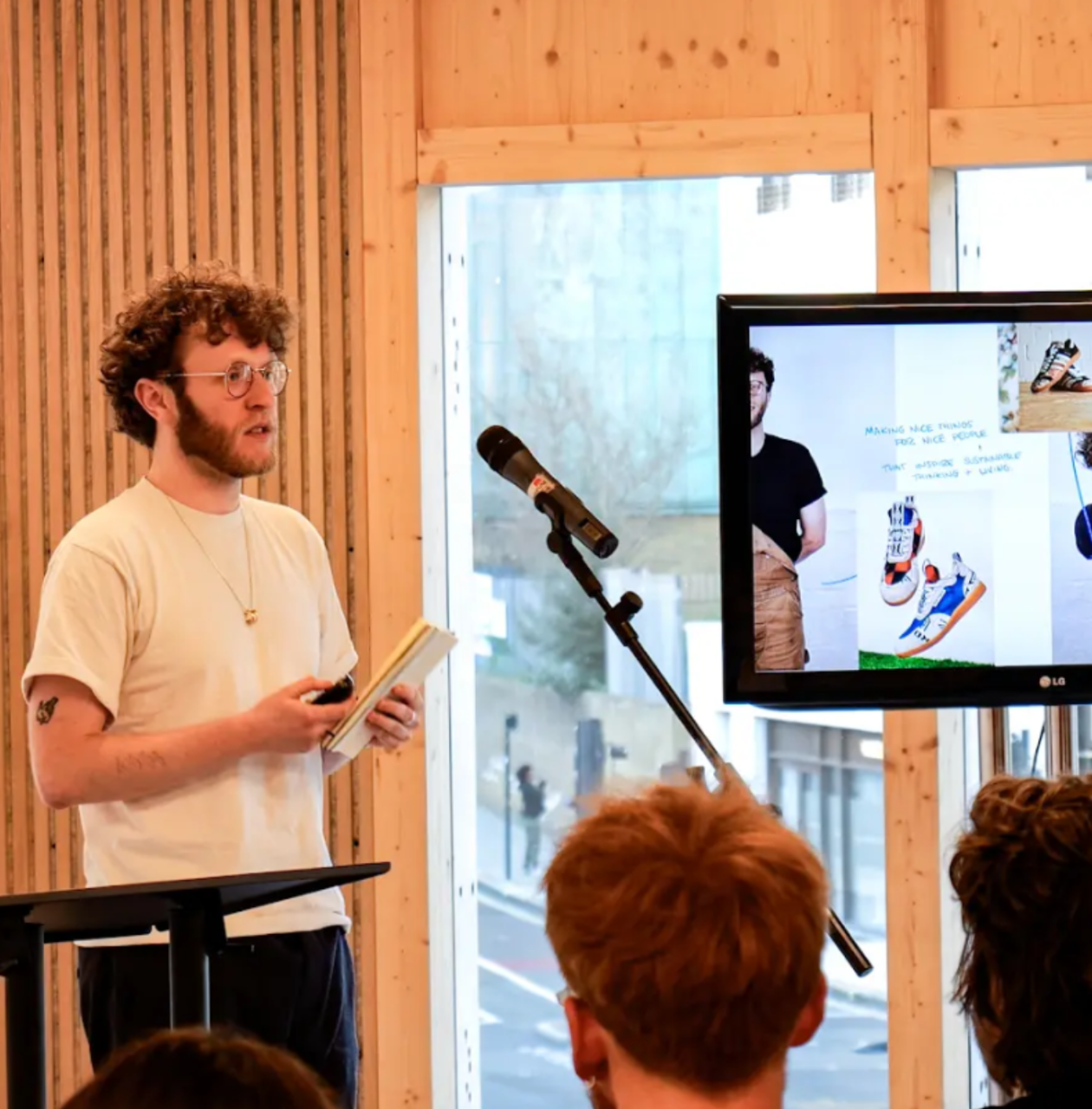 A man gives a presentation to an audience, standing beside a microphone and a large screen displaying images of sneakers and a design process.