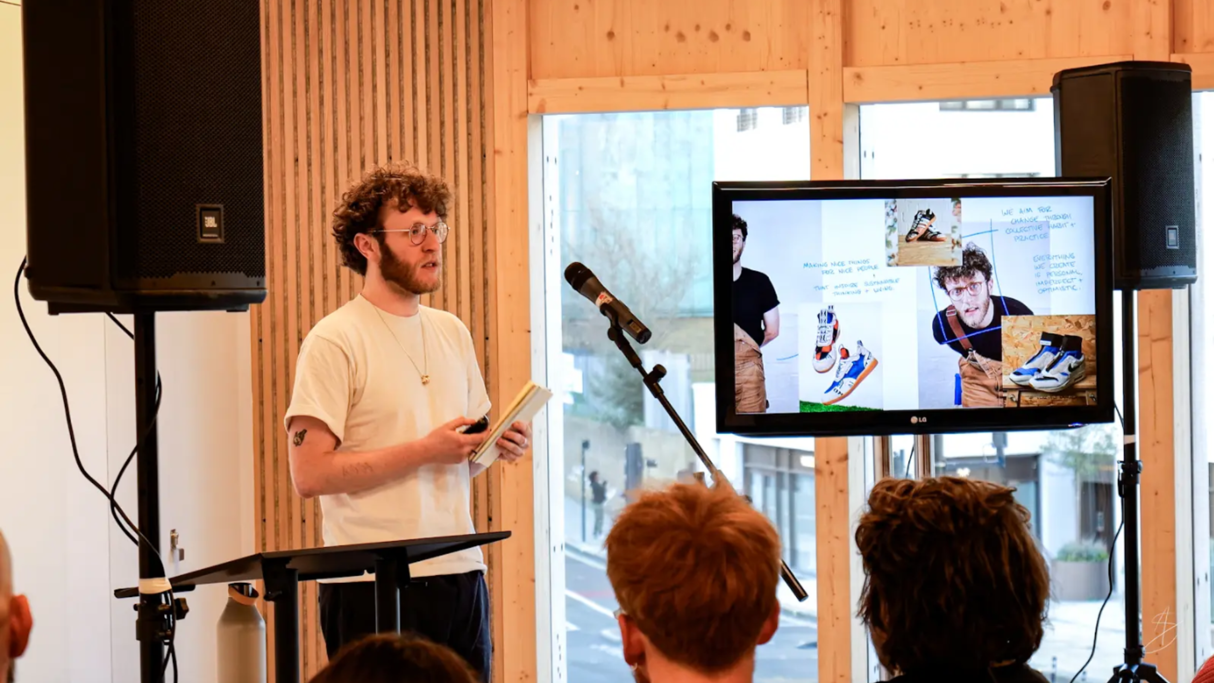 A man gives a presentation to an audience, standing beside a microphone and a large screen displaying images of sneakers and a design process.