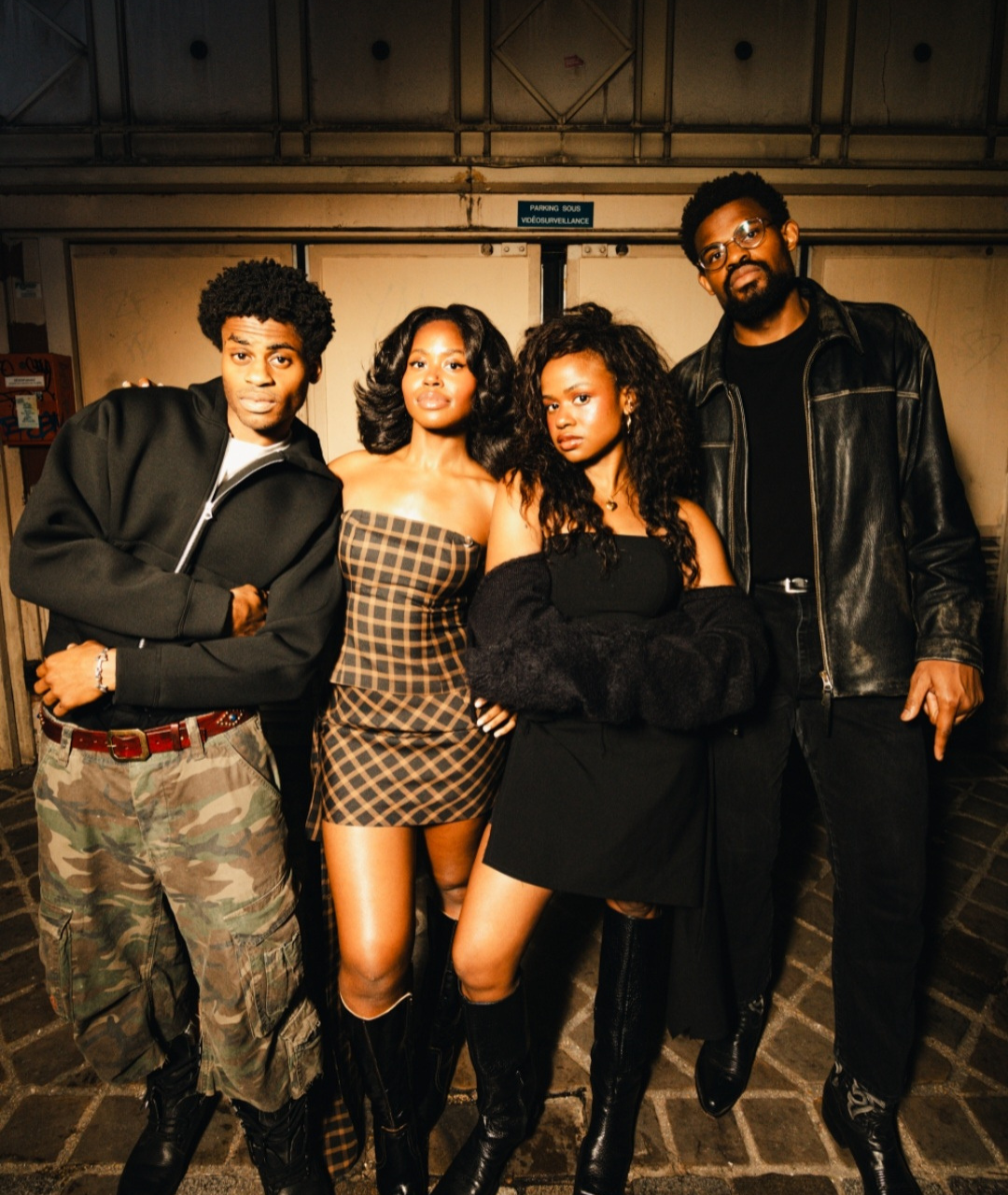 Four members of the band Infinity Song pose confidently together in coordinated stylish outfits against an industrial backdrop.