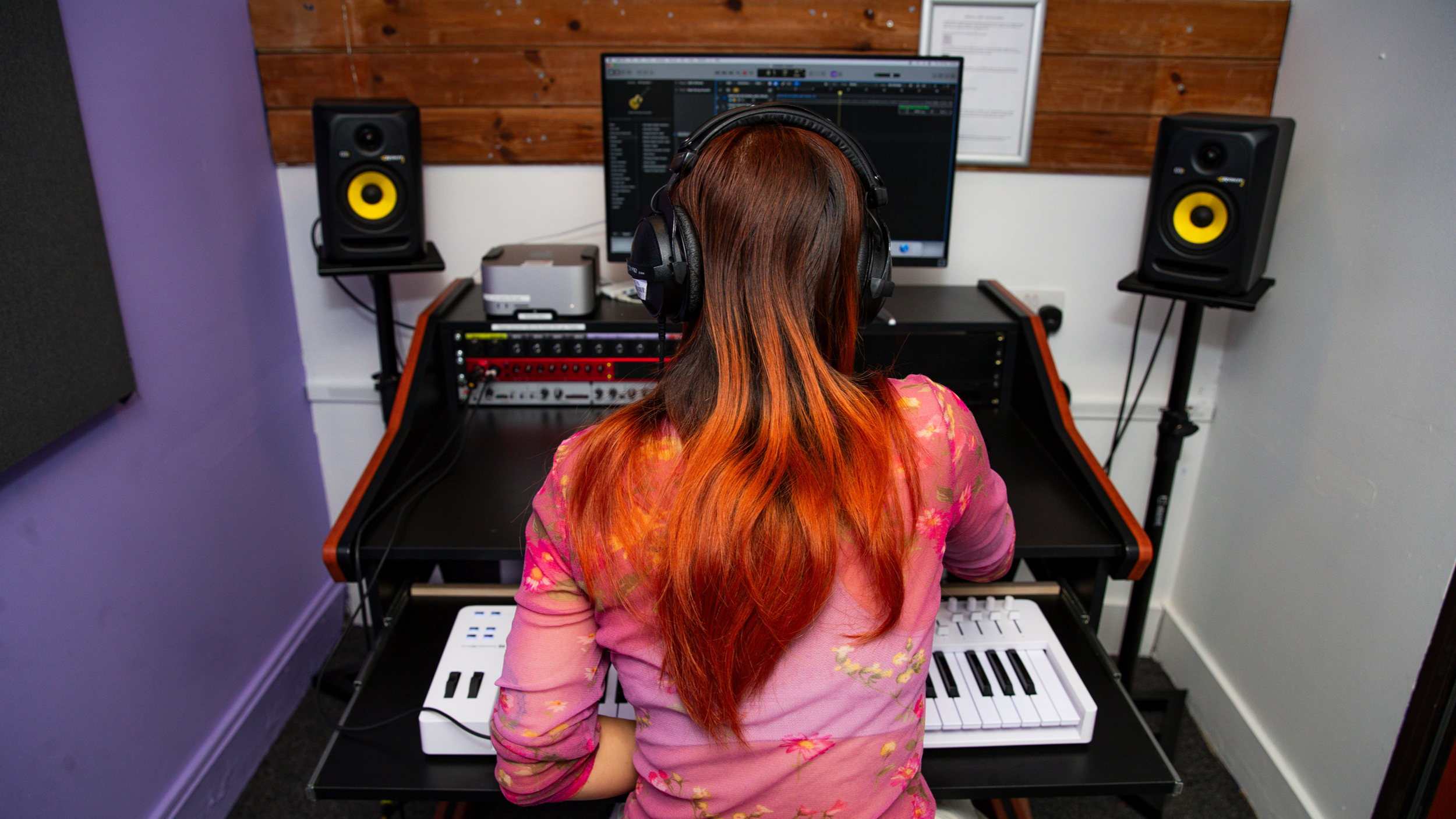A person with long, dark red and orange ombre hair is sitting at a music production workstation