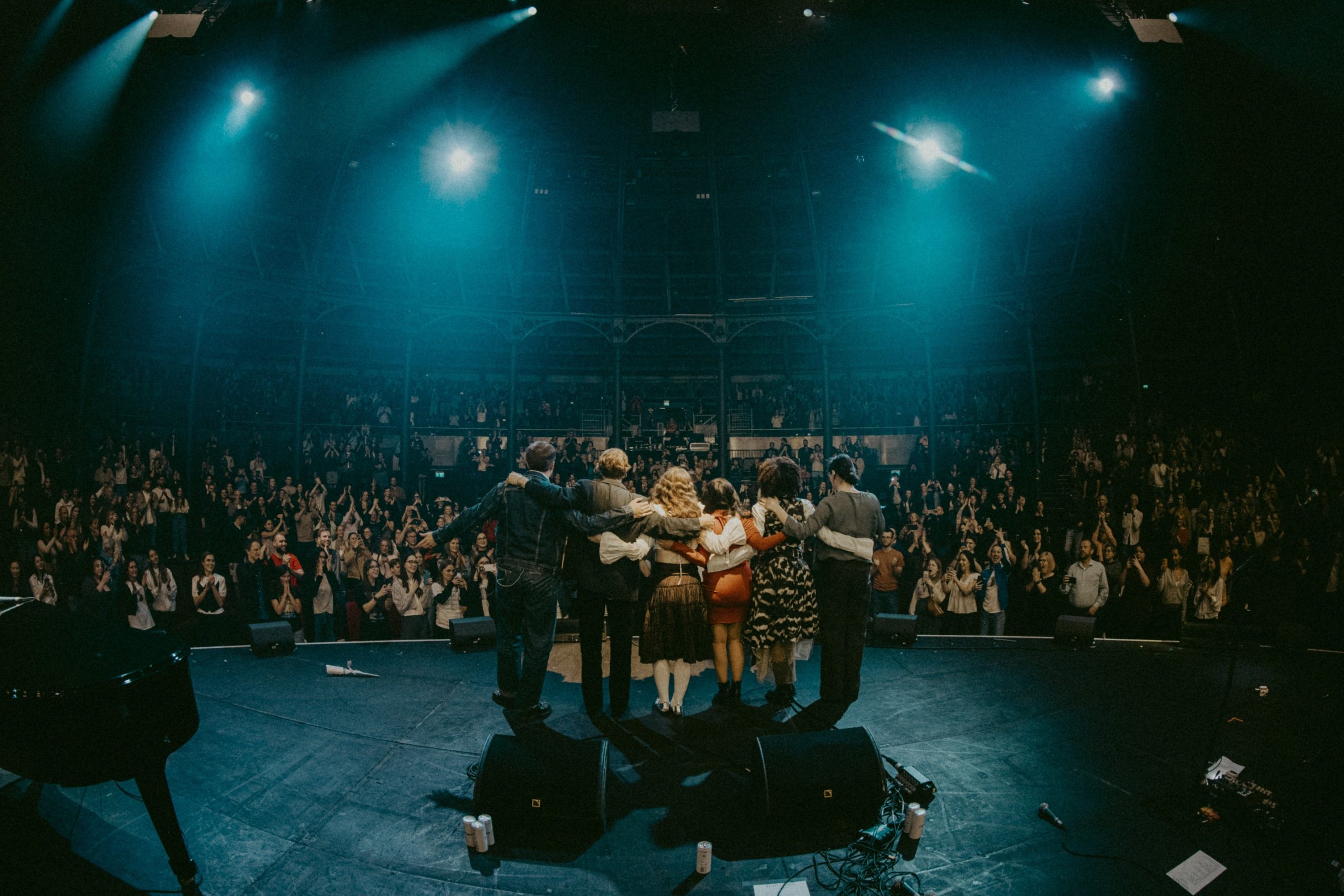 Six people stand arm in arm, facing out on a stage. A large audience applauds them under big blue stage lights.