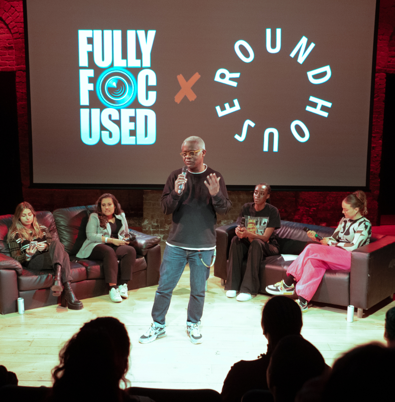 A speaker holds a microphone addressing an audience in front of a large screen displaying 