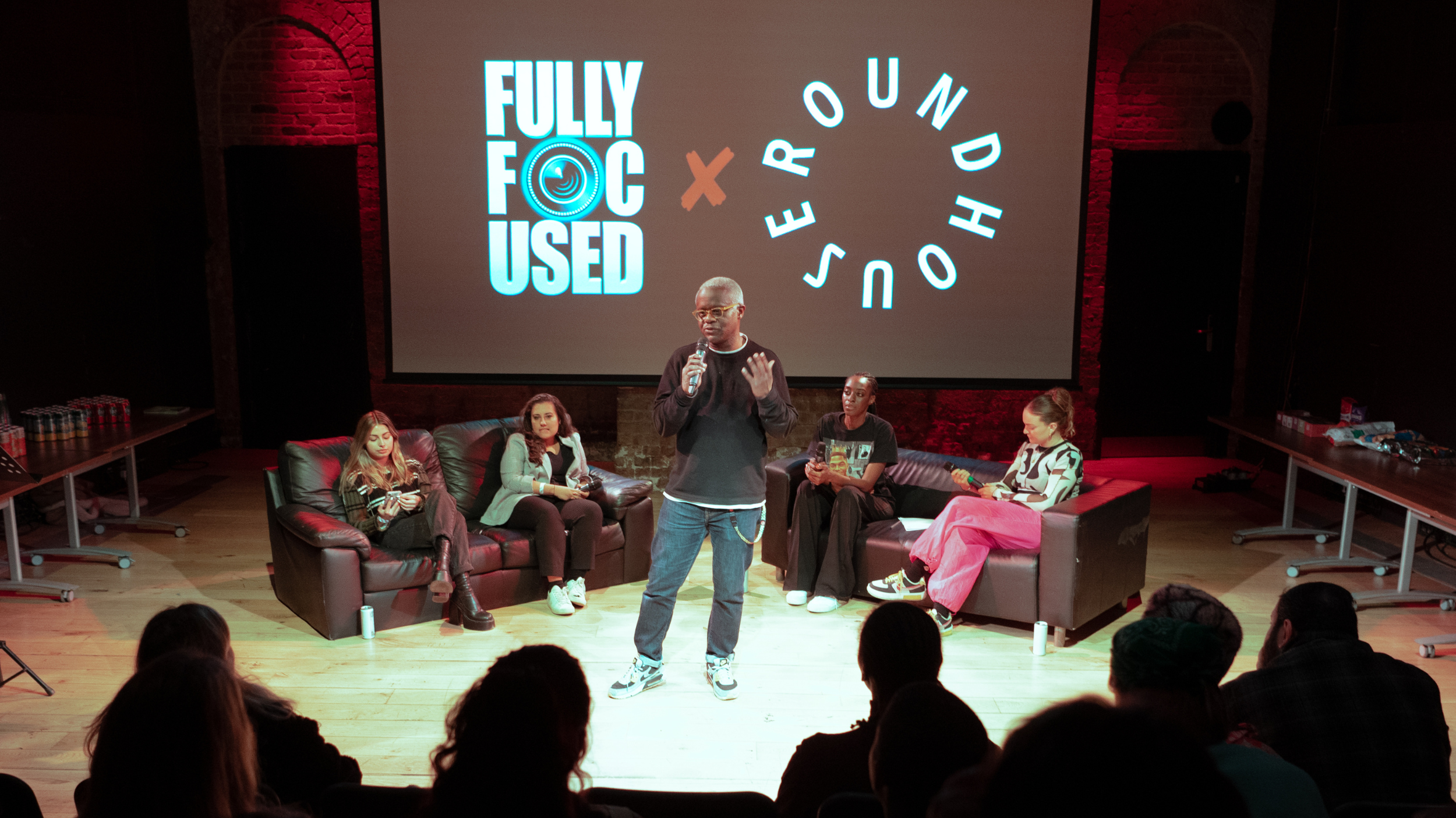 A speaker holds a microphone addressing an audience in front of a large screen displaying 